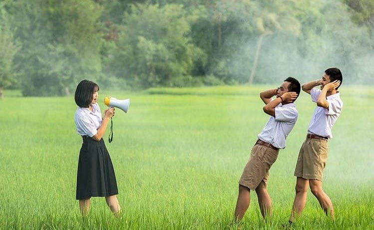 chinese woman using a megaphone sayong something to two chinese men holding close their ears
