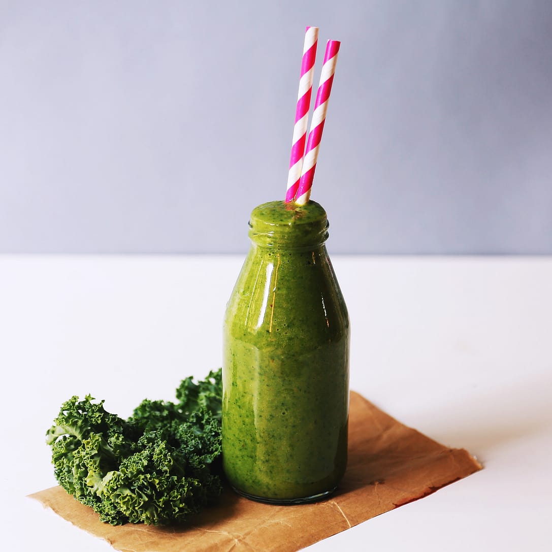 green juice in a bottle with 2 straws, a bunch of kale beside the bottle